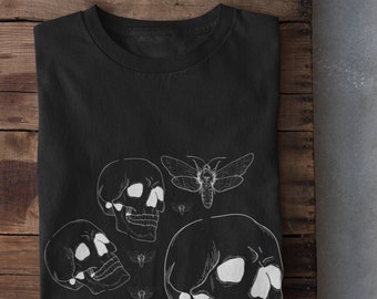 Puede incluir: Camiseta negra con un diseño gráfico blanco de calaveras y polillas. El diseño presenta tres calaveras y una polilla con las alas extendidas.