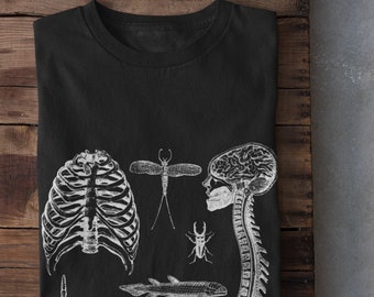 Puede incluir: Camiseta negra con ilustraciones anatómicas blancas de una caja torácica, insecto, cerebro, columna vertebral y pez. La camiseta está doblada sobre una superficie de madera.