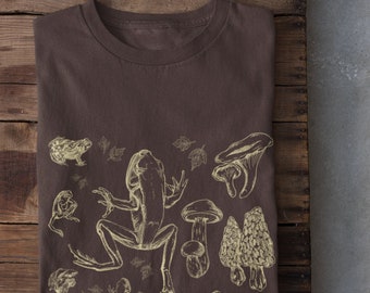 Può includere: Una maglietta marrone con un disegno giallo chiaro di una rana, funghi e foglie.
