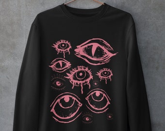 May include: Black sweatshirt featuring a pink graphic print of various eyes. The eyes are depicted in a stylised, slightly eerie design.