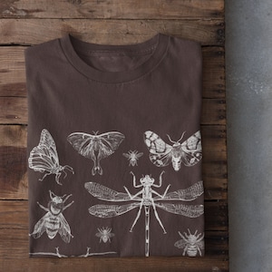 May include: A brown t-shirt with a white print of various insects, including butterflies, moths, bees, and a dragonfly. The shirt is folded and laid on a wooden surface.
