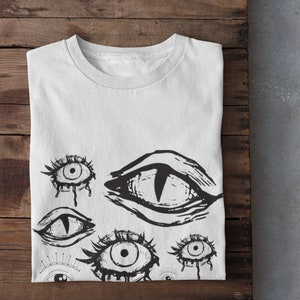 May include: White t-shirt with a black and white graphic print of multiple stylized eyes. The eyes are drawn in a creepy, abstract style.
