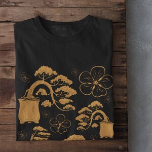 Może przedstawiać: Czarny t-shirt z złotym wzorem bonsai i kwiatów.