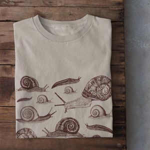 May include: A beige t-shirt featuring a brown snail design. The design includes several snails of varying sizes and shapes.