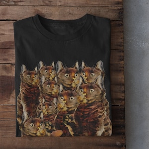 Puede incluir: Camiseta negra con un estampado de varios gatos tabby marrones mirando al espectador.