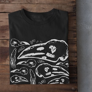 Puede incluir: Camiseta negra con un diseño gráfico blanco de calaveras de pájaros y formas abstractas. La camiseta está doblada sobre una superficie de madera, con zapatillas blancas visibles en la parte inferior. Una prenda informal de inspiración gótica.
