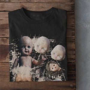 Può includere: Maglietta nera con una stampa grafica di diversi volti di bambole. I volti sono usurati e sbiaditi, dando al design un aspetto vintage.