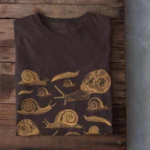 Könnte beinhalten: Ein braunes T-Shirt mit einem goldenen Druck verschiedener Schnecken und Nacktschnecken.