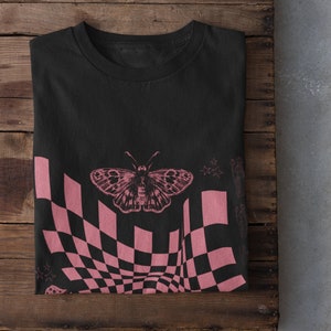May include: Black t-shirt with a pink checkered pattern and a butterfly graphic. The pattern is a distorted checkerboard with a pink butterfly in the center.