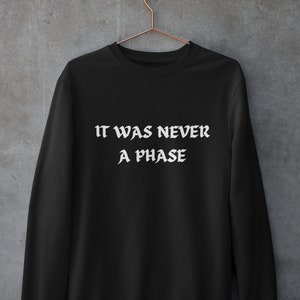 Pode incluir: Camisola preta com o texto branco "IT WAS NEVER A PHASE". A peça de roupa está pendurada num cabide cor de cobre contra uma parede cinzenta. O texto está numa fonte gótica.
