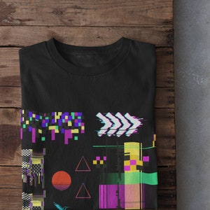 Puede incluir: Una camiseta negra con un diseño geométrico colorido que presenta cuadrados pixelados, triángulos y flechas. El diseño tiene un efecto de glitch, creando una estética retro y digital.