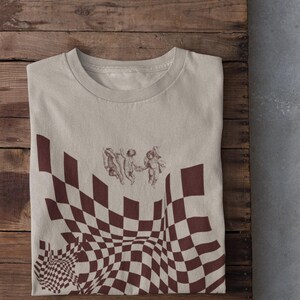 May include: A beige t-shirt featuring a brown and white checkered pattern with a small image of three cherubs in the top centre of the shirt.
