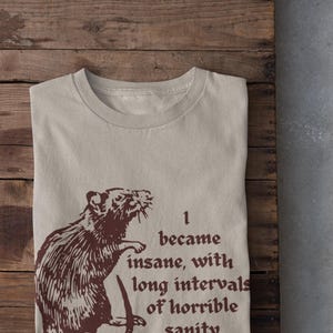 Peut inclure: T-shirt beige avec une illustration de rat marron et le texte "I became insane, with long intervals of horrible sanity." Le t-shirt est plié sur une surface en bois, avec des baskets blanches visibles.