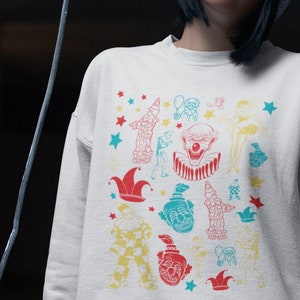 May include: A white sweatshirt with a colorful pattern of clowns, stars, and other circus-themed imagery. The sweatshirt has a crew neck and long sleeves.