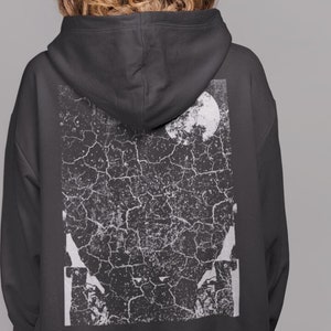 Może przedstawiać: Czarna bluza z kapturem z dużym, prostokątnym nadrukiem na plecach. Grafika to postarzany, spękany wzór w kolorze białym i szarym, z pełnią księżyca i abstrakcyjnymi kształtami. Bluza ma kaptur i luźny krój.