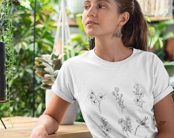 May include: A woman wearing a white t-shirt with a black line drawing of flowers on it. She is sitting on a wooden bench in a greenhouse setting.