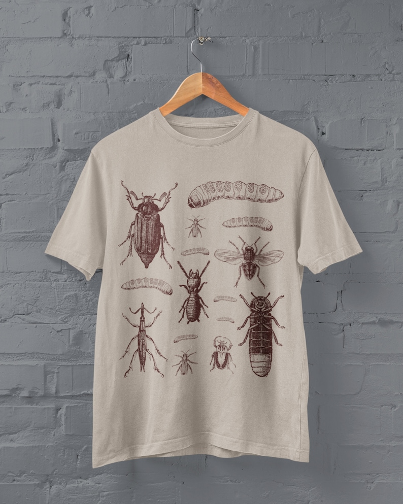 May include: Beige t-shirt with a brown illustration of insects, such as beetles, ants, and a caterpillar. The graphic is arranged in a grid. The shirt hangs on a wooden hanger against a grey brick wall.