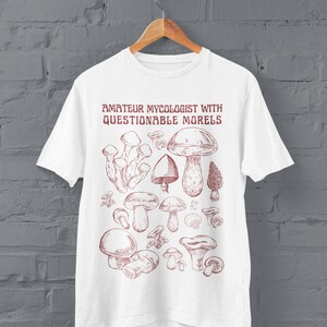 Może przedstawiać: Biała koszulka z czerwonym nadrukiem r&oacute;żnych grzyb&oacute;w i tekstem "Amateur Mycologist with Questionable Morels".