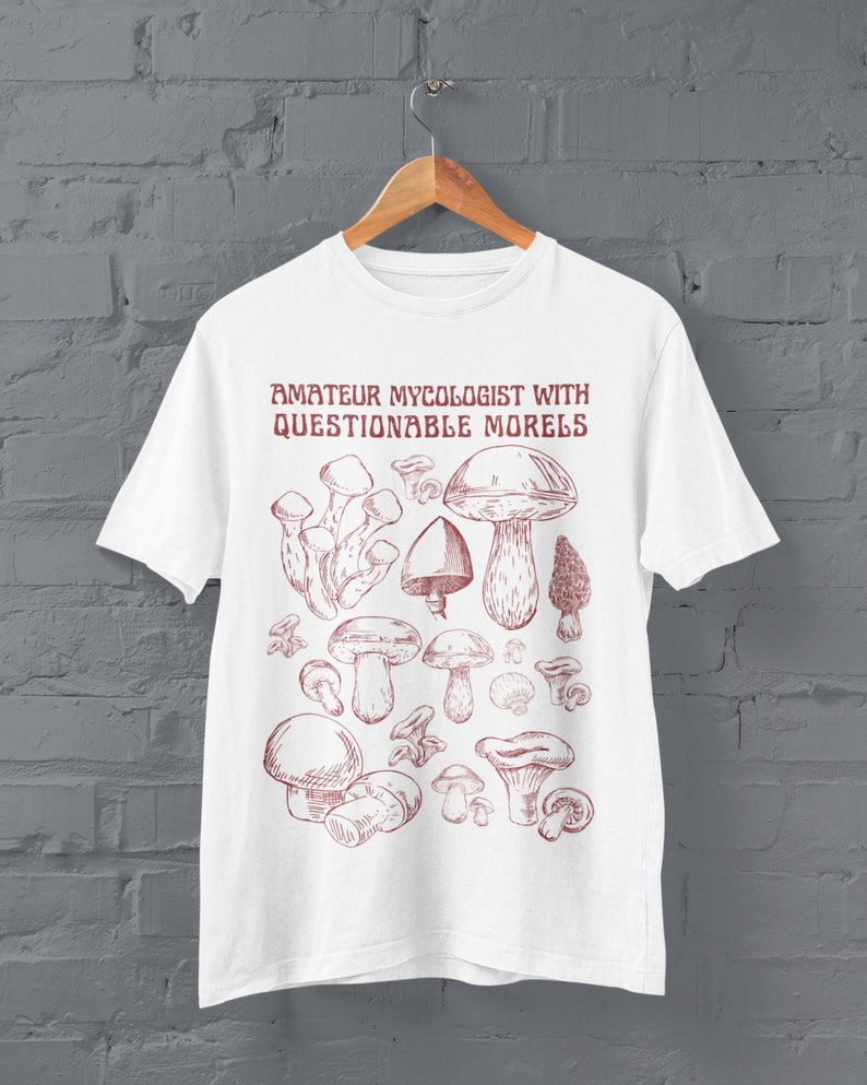 Może przedstawiać: Biała koszulka z czerwonym nadrukiem r&oacute;żnych grzyb&oacute;w i tekstem "Amateur Mycologist with Questionable Morels".