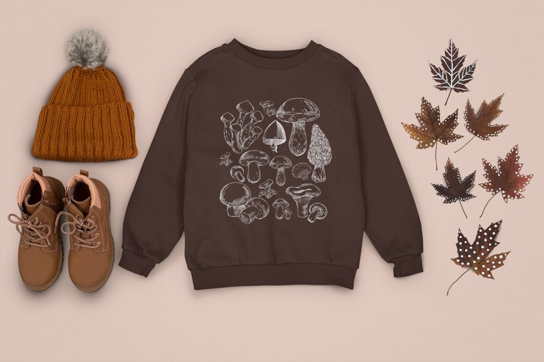 Puede incluir: Una sudadera marr&oacute;n con un estampado de setas blancas. La sudadera tiene mangas largas y cuello redondo. Tambi&eacute;n hay botas marrones, un gorro de punto marr&oacute;n con un pomp&oacute;n blanco y hojas marrones con lunares blancos.