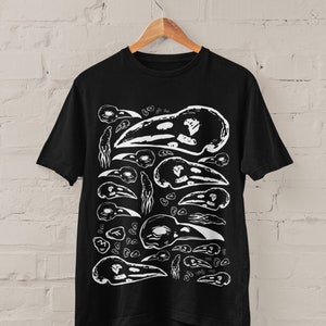 Op de afbeelding: Zwart T-shirt met een wit grafisch patroon van vogel schedels. Het ontwerp bevat illustraties van vogel schedels in verschillende maten en posities. Het T-shirt hangt aan een houten hanger tegen een witte bakstenen muur.