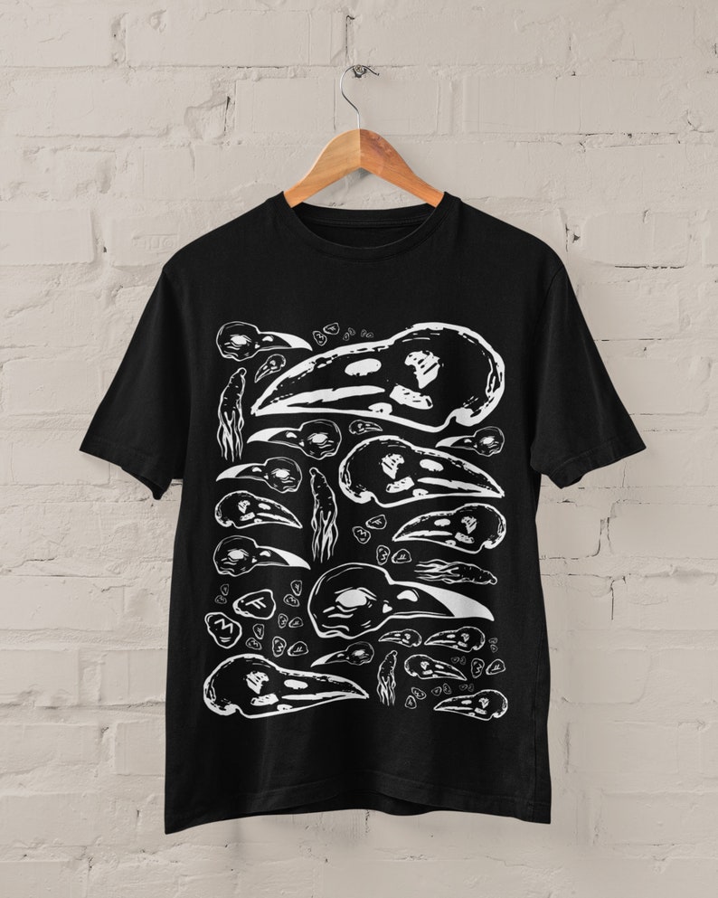 Op de afbeelding: Zwart T-shirt met een wit grafisch patroon van vogel schedels. Het ontwerp bevat illustraties van vogel schedels in verschillende maten en posities. Het T-shirt hangt aan een houten hanger tegen een witte bakstenen muur.