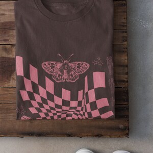 May include: A brown t-shirt with a pink checkered pattern, a butterfly, and stars. The shirt is folded and lying on a wooden surface.