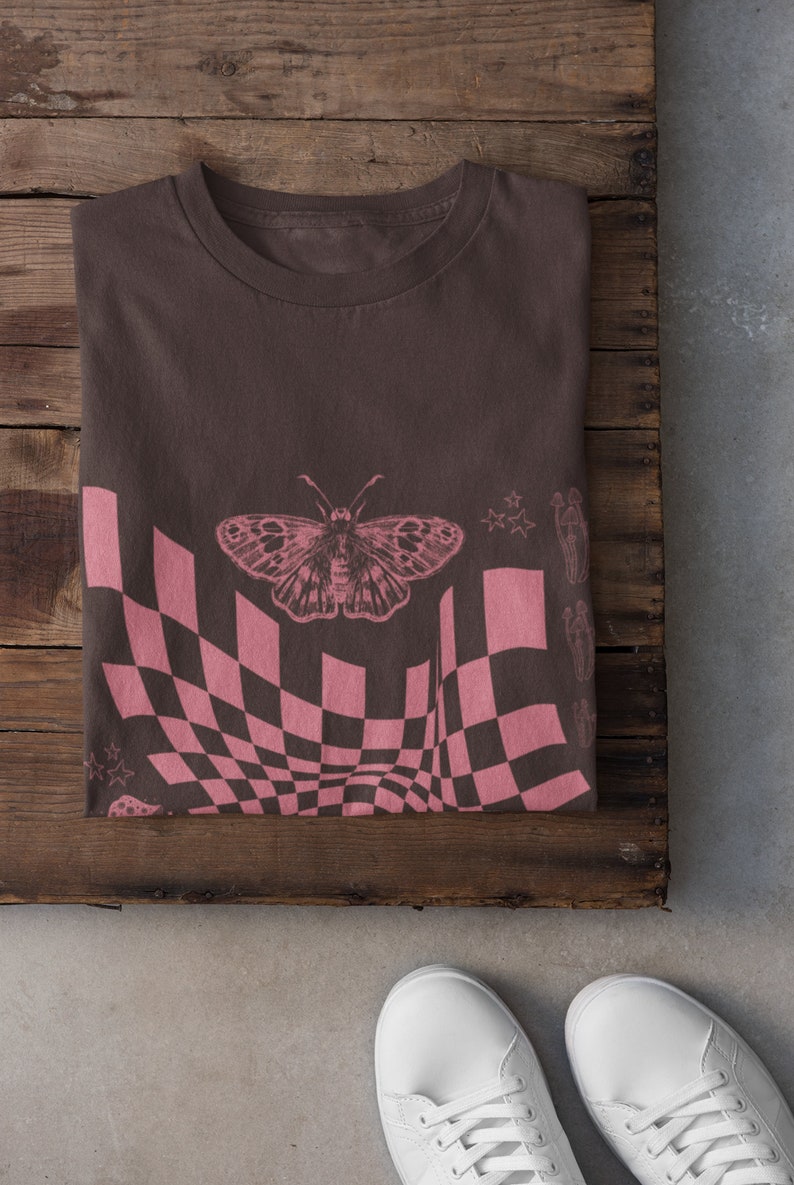 May include: A brown t-shirt with a pink checkered pattern, a butterfly, and stars. The shirt is folded and lying on a wooden surface.