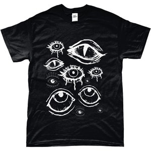 May include: Black t-shirt featuring a white graphic of various stylized eyes. The eyes have different shapes and sizes, some with teardrop details. The design is printed on a black, short-sleeved t-shirt.