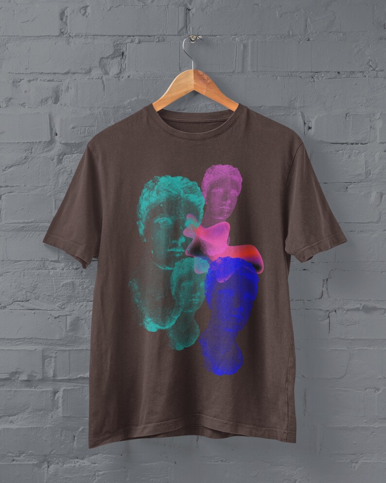 May include: A brown t-shirt with a graphic print of three colorful, abstract faces. The faces are in shades of blue, green, and pink.