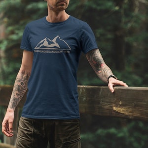 Puede incluir: Una camiseta azul marino con un gráfico de montaña blanco y el texto "Two Hundred And Eighty Two" impreso en la parte delantera.