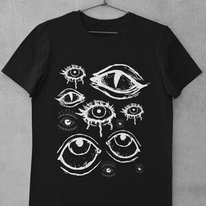 May include: Black t-shirt featuring a white graphic of various stylized eyes. The eyes have different shapes and sizes, with some appearing to be crying. The design is a bold, artistic statement, perfect for those who appreciate unique apparel.