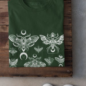 May include: A dark green t-shirt with a white print of moths and crescent moons. The moths are stylized and have intricate details. The print is repeated across the bottom of the shirt.