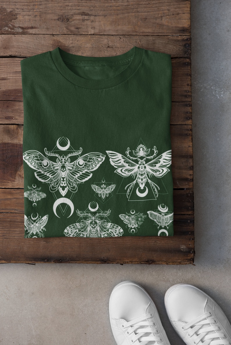 May include: A dark green t-shirt with a white print of moths and crescent moons. The moths are stylized and have intricate details. The print is repeated across the bottom of the shirt.