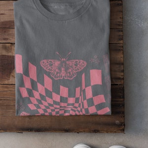 May include: A gray t-shirt with a pink checkered pattern and a butterfly graphic. The pattern is distorted, creating a wavy effect.