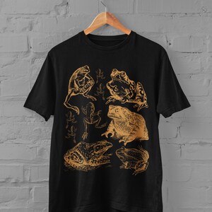 Pode incluir: Camiseta preta com uma estampa dourada de v&aacute;rios sapos e r&atilde;s. A estampa apresenta uma variedade de poses de sapos, incluindo sentados, pulando e nadando.