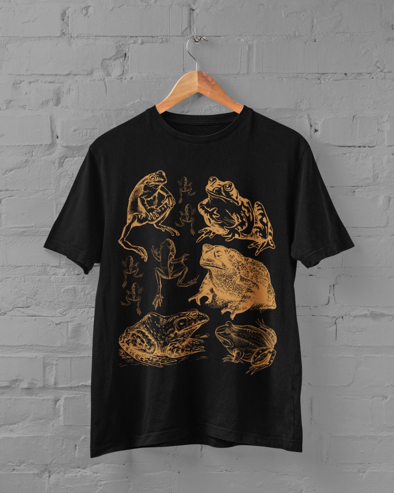 Pode incluir: Camiseta preta com uma estampa dourada de v&aacute;rios sapos e r&atilde;s. A estampa apresenta uma variedade de poses de sapos, incluindo sentados, pulando e nadando.