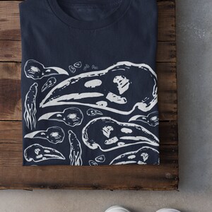 Op de afbeelding: Marineblauwe t-shirt met een witte grafische print van vogel schedels en abstracte vormen. Het gevouwen shirt ligt op een houten oppervlak, met witte sneakers op de voorgrond. Het ontwerp wordt herhaald over het shirt.