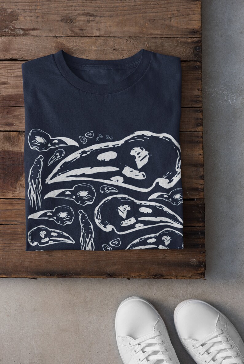 Op de afbeelding: Marineblauwe t-shirt met een witte grafische print van vogel schedels en abstracte vormen. Het gevouwen shirt ligt op een houten oppervlak, met witte sneakers op de voorgrond. Het ontwerp wordt herhaald over het shirt.