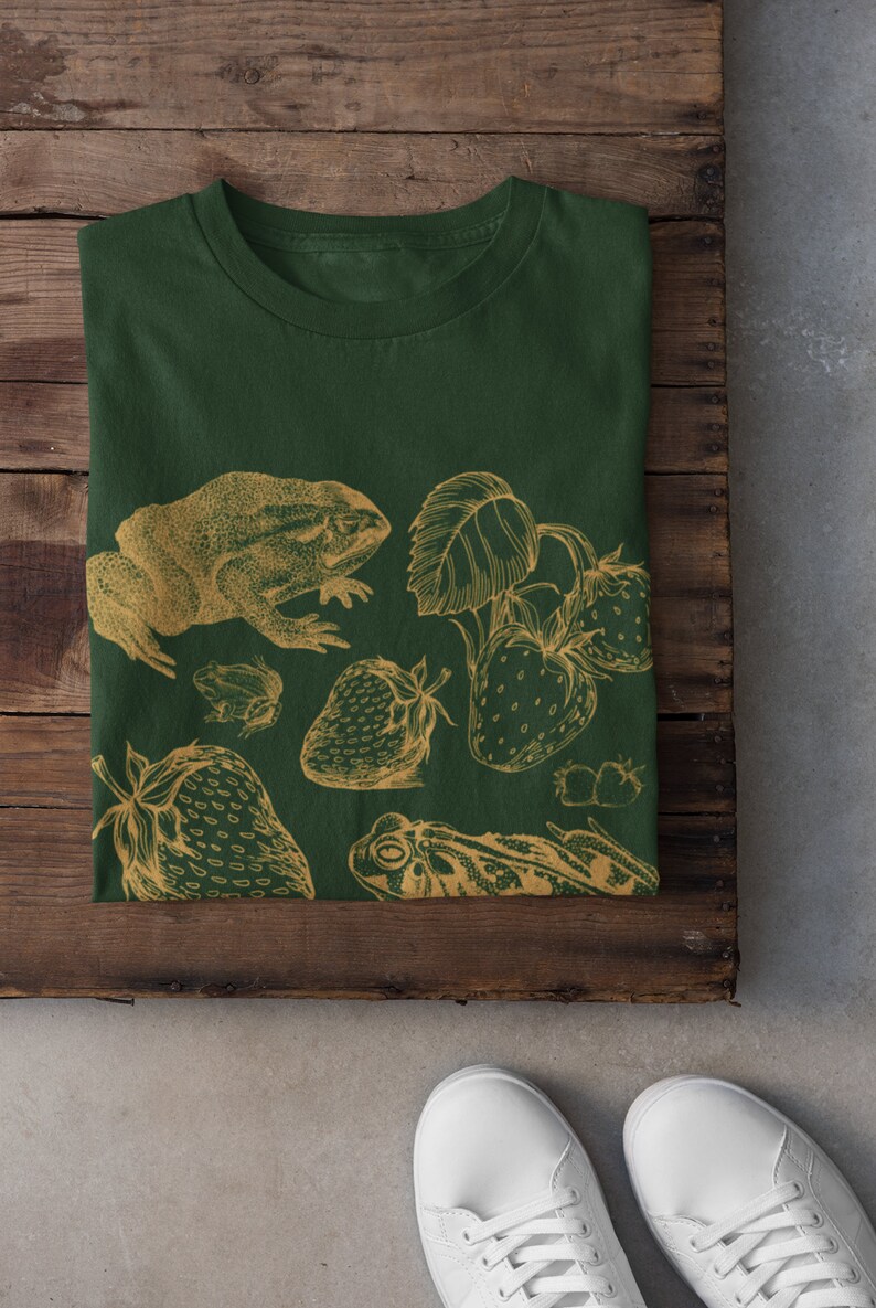 Pu&ograve; includere: Una maglietta verde con un design a contorno dorato di rane, fragole e foglie.