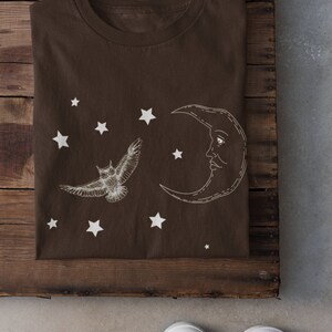 May include: A brown t-shirt with a white crescent moon and a white owl flying in front of it. There are white stars scattered around the moon and owl. The t-shirt is folded and lying on a wooden surface.