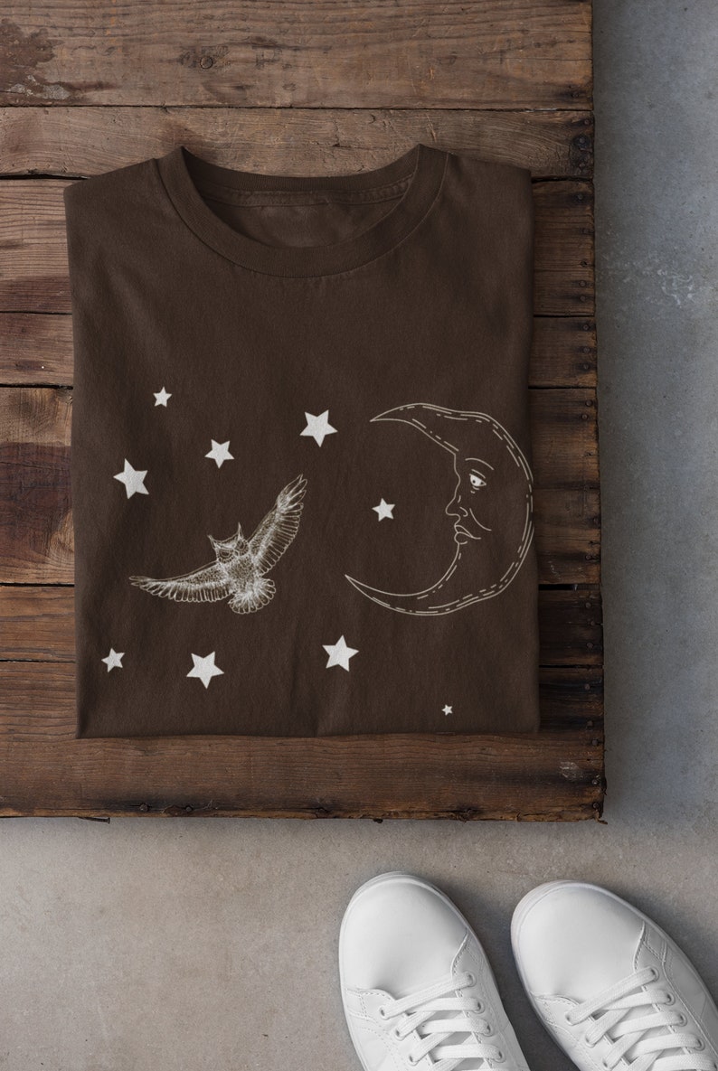 May include: A brown t-shirt with a white crescent moon and a white owl flying in front of it. There are white stars scattered around the moon and owl. The t-shirt is folded and lying on a wooden surface.