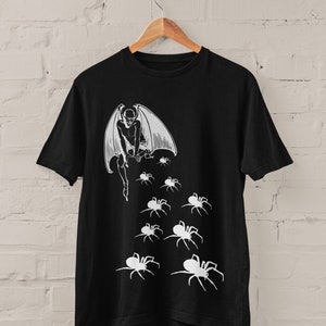 Op de afbeelding: Zwart T-shirt met een witte grafische print van een gevleugelde demon die op een stapel spinnen zit. De demon heeft een lange staart en wordt omringd door acht spinnen.