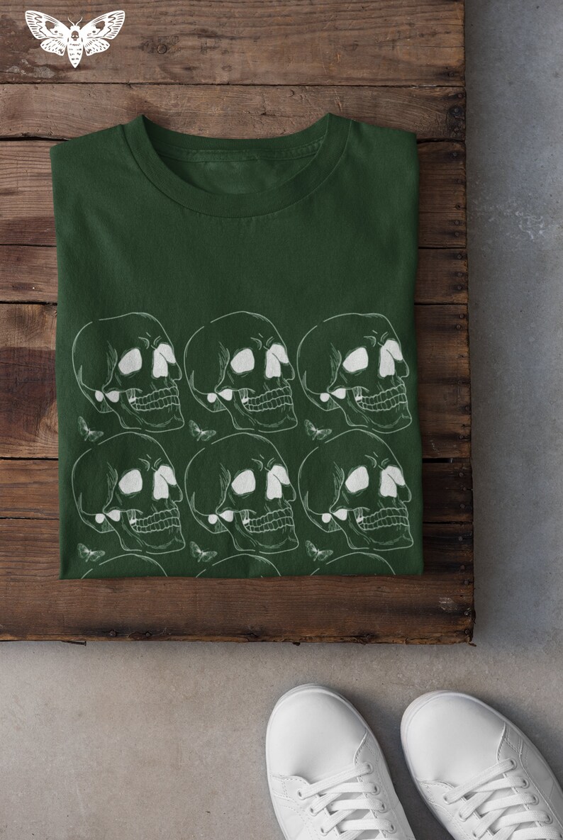 May include: A dark green t-shirt with a white skull and moth pattern. The shirt is folded and laid on a wooden surface.