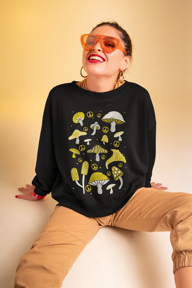 Puede incluir: Sudadera negra con un estampado gr&aacute;fico de setas amarillas y blancas. El estampado presenta varios tipos de setas con s&iacute;mbolos de paz.