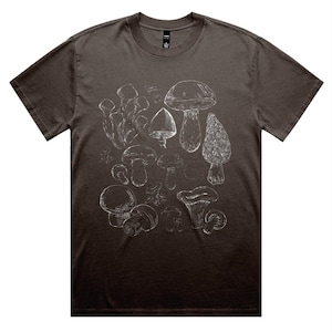 Puede incluir: Una camiseta marrón oscuro con una ilustración blanca de varias setas. El diseño incluye dibujos detallados de diferentes tipos de setas, creando un tema botánico. La camiseta tiene un cuello redondo clásico y mangas cortas.