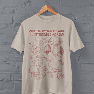Może przedstawiać: Beżowa koszulka z czerwonym nadrukiem r&oacute;żnych grzyb&oacute;w i tekstem "Amateur Mycologist with Questionable Morels".
