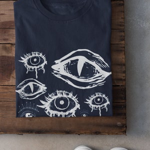 May include: Navy blue t-shirt featuring a white eye design. The graphic includes various stylized eye illustrations with different shapes and details. The shirt is folded on a wooden surface, with white sneakers visible below.