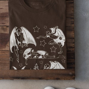 May include: A brown t-shirt with a white print of multiple winged figures and stars. The figures are in various poses, some flying, some crawling, and some standing.