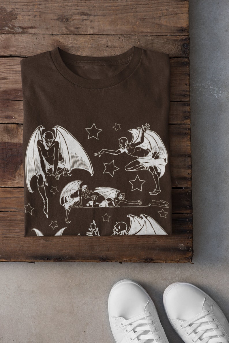 May include: A brown t-shirt with a white print of multiple winged figures and stars. The figures are in various poses, some flying, some crawling, and some standing.